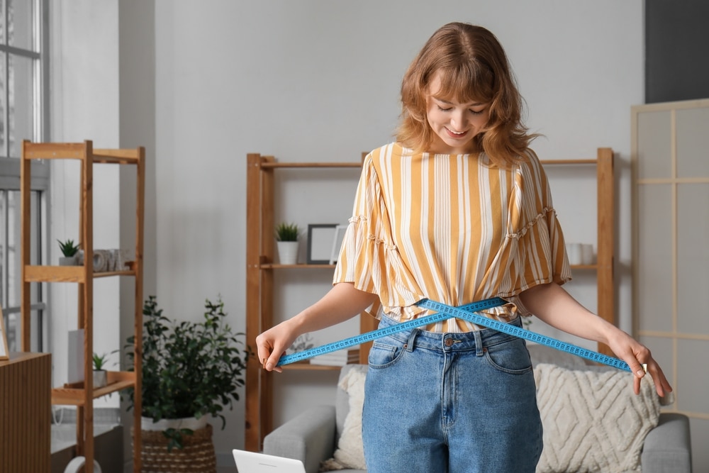 Redhead woman in a bedroom wrapping a tape measure around her waist.